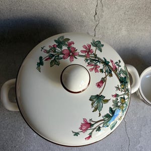 Peut inclure: Plat &agrave; four en c&eacute;ramique cr&egrave;me avec couvercle. Le couvercle pr&eacute;sente un motif floral avec des fleurs roses et des feuilles vertes. Le plat a deux poign&eacute;es et un bouton rond. Mesures en cm.