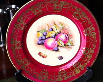 May include: A red and gold decorative plate with a white center featuring a painted image of fruit, including grapes, plums, and berries.