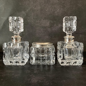 May include: Three clear glass bottles with silver tops. The bottles are square shaped and have a textured design. The bottles are arranged in a row on a dark background.