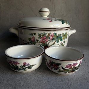 Peut inclure: Un plat de service en c&eacute;ramique blanche avec un couvercle et deux bols assortis. L'ensemble pr&eacute;sente un motif floral avec des fleurs et des feuilles roses et vertes. Les bords sont bord&eacute;s d'une bordure marron fonc&eacute;. Le plat de service a deux poign&eacute;es.
