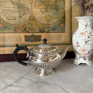 Könnte beinhalten: Eine silberne Teekanne mit schwarzem Griff und dekorativem Deckel. Die Teekanne hat ein geripptes Design am unteren Teil und steht auf einem kleinen Sockel. Eine Vintage-Karte und eine Blumenvase befinden sich im Hintergrund.