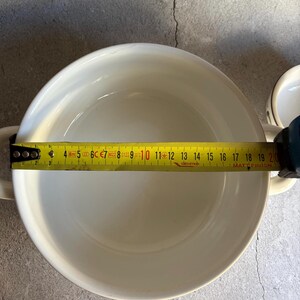 Peut inclure: Un bol en c&eacute;ramique blanc avec deux poign&eacute;es, mesur&eacute; avec un m&egrave;tre ruban jaune. Le diam&egrave;tre du bol est d'environ 20 cm. Le m&egrave;tre ruban affiche la mesure en centim&egrave;tres. Le bol est sur une surface grise.