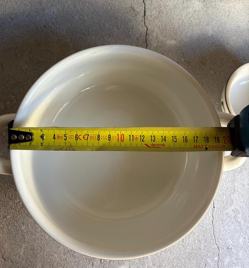 Peut inclure: Un bol en c&eacute;ramique blanc avec deux poign&eacute;es, mesur&eacute; avec un m&egrave;tre ruban jaune. Le diam&egrave;tre du bol est d'environ 20 cm. Le m&egrave;tre ruban affiche la mesure en centim&egrave;tres. Le bol est sur une surface grise.