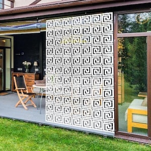 May include: A white geometric privacy screen with a Greek key design is installed on a patio. The screen is made of squares. The patio has a table and chairs. The house has a brown roof and is surrounded by a green lawn.