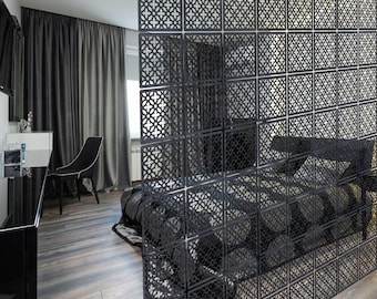 May include: A black decorative room divider with a geometric pattern, separating a bedroom area. The divider is made of square panels with a repeating openwork design. The room features a bed, chair, and dark curtains, creating a modern, monochromatic aesthetic.