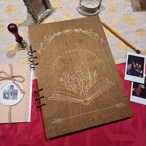 Pode incluir: Um diário de madeira com encadernação em espiral. A capa é decorada com um desenho gravado de um livro com flores e o texto "Книга наших пригод".