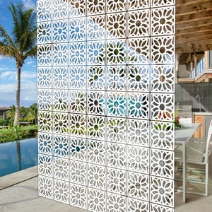May include: A white decorative screen made of multiple square panels. Each panel has a floral, openwork pattern. The screen is positioned outside, with a view of a pool, palm trees, and a modern house visible through the openings.