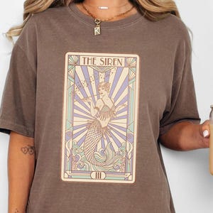 May include: Espresso-colored t-shirt featuring a tarot card design with the text "THE SIREN." The card depicts a mermaid with a vintage art style. The shirt is worn with denim shorts.