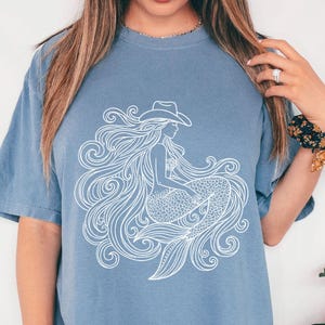 May include: A blue jean colored t-shirt featuring a white graphic of a mermaid wearing a cowboy hat. The mermaid has long flowing hair and a fishtail. The shirt is a comfortable, casual style.