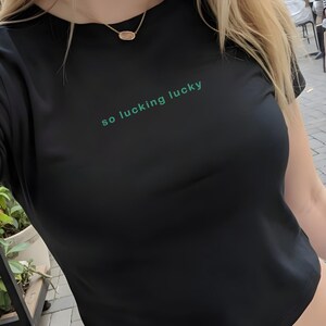 May include: Black crop top with the text "so fucking lucky" in green. The shirt is made of a soft material and has a crew neck. The model is wearing blue jeans and a gold necklace.