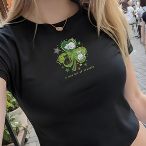 May include: Black crop top with a shamrock design featuring three cats and the text "a wee bit of trouble". The shamrock is green with white, black, and green cats. The shirt is made of a soft, dark material.