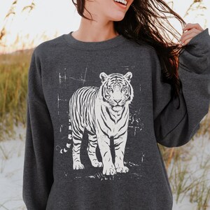 May include: Dark heather gray sweatshirt featuring a white tiger graphic. The tiger is standing with its front paws forward. The text "DARK HEATHER" is at the bottom right corner of the image. The sweatshirt has long sleeves.