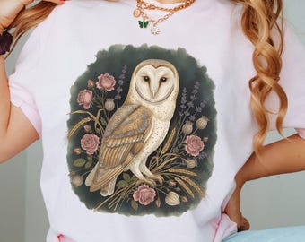 May include: A pale pink t-shirt with a detailed illustration of a barn owl, pink roses, wheat, and lavender. The owl has a white face and brown feathers. The word "BLOSSOM" is printed in the bottom right corner of the image.