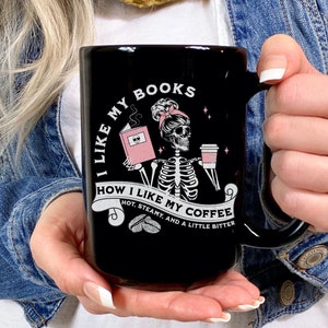 May include: Black ceramic mug with a humorous design. The mug features a skeleton reading a book and holding coffee, with the text "I LIKE MY BOOKS, HOW I LIKE MY COFFEE, HOT, STEAMY, AND A LITTLE BITTER."