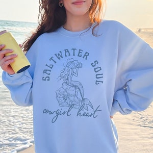 woman wearing a light blue sweatshirt with a blue design of a mermaid wearing a cowgirl hat and the phrase saltwater soul cowgirl heart