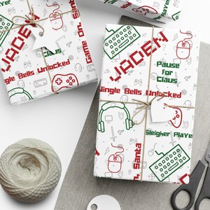 Peut inclure: Papier d'emballage blanc avec un motif sur le thème du jeu vidéo rouge et vert. Le papier présente un motif répétitif de manettes de jeu vidéo, de joysticks, de casques d'écoute et d'autres icônes de jeu. Le texte "JADEN" est répété dans tout le motif. Le texte "Jingle Bells Unlocked" et "Pause for Claus" sont également inclus dans le motif.
