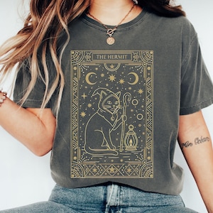 May include: A dark gray t-shirt featuring a gold-colored illustration of a cat in a hooded robe, holding a staff and lantern. The text "THE HERMIT" is at the top of the design, with crescent moons and stars. The shirt is a Pepper color.