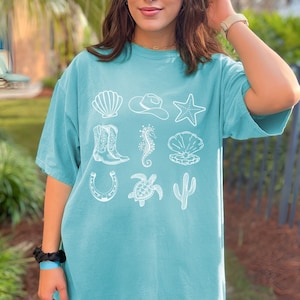 woman wearing a seafoam shirt with rows of cowgirl and coastal elements.
