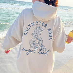 May include: A cream-coloured hoodie with a mermaid graphic on the back. The graphic says "Saltwater Soul" and "Cowgirl Heart".
