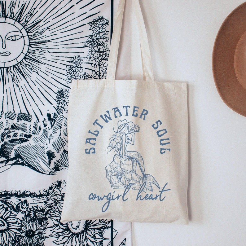 beige canvas tote bag with a blue design showing a mermaid cowgirl sitting on a rock and the phrase saltwater soul cowgirl heart