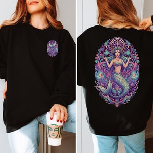 May include: Black sweatshirt with a mermaid design. The front features a small, oval-shaped mermaid tail design in purple and blue. The back displays a larger, detailed illustration of a mermaid with flowing hair, a crown, and a teal tail, surrounded by floral elements.