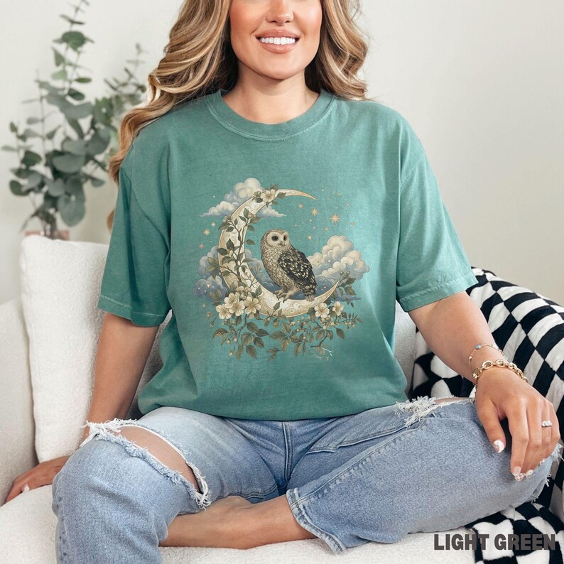 May include: Light green t-shirt featuring a detailed illustration of an owl perched on a crescent moon, surrounded by clouds and flowers. The text "LIGHT GREEN" is at the bottom right corner. The shirt is a relaxed fit.