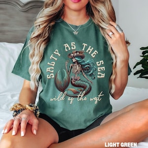May include: Light green t-shirt featuring a skeleton mermaid graphic with a cowboy hat and the text "Salty as the Sea, Wild as the West". The mermaid has blue hair and a brown tail.