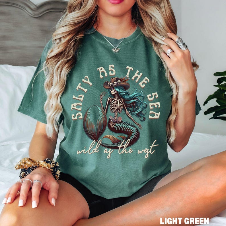 May include: Light green t-shirt featuring a skeleton mermaid graphic with a cowboy hat and the text "Salty as the Sea, Wild as the West". The mermaid has blue hair and a brown tail.