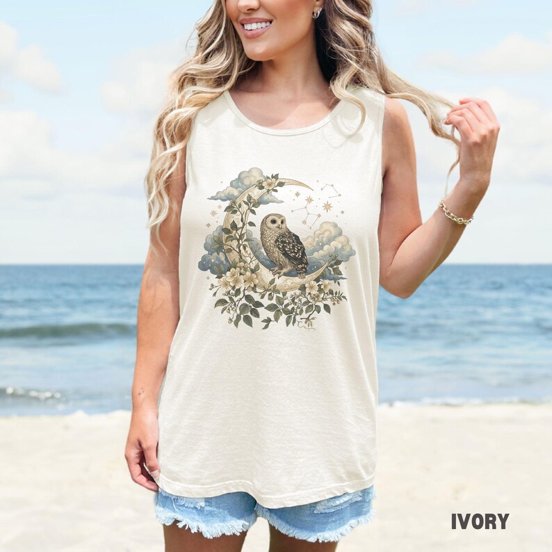 May include: Ivory-colored tank top featuring a detailed illustration of an owl perched on a crescent moon, surrounded by clouds, flowers, and stars. The text "IVORY" is printed at the bottom. The top is sleeveless.