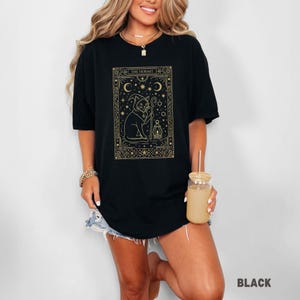 May include: Black t-shirt featuring a gold illustration of a cat in a hooded cloak holding a lantern and staff, with the text "THE HERMIT" at the top. The design is framed by stars, moons, and geometric patterns. The word "BLACK" is at the bottom right.