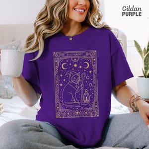 May include: A purple t-shirt featuring a gold illustration of a cat in a hooded cloak holding a staff and lantern, with the text "THE HERMIT". The design includes stars, moons, and geometric borders. The Gildan Purple brand is visible.