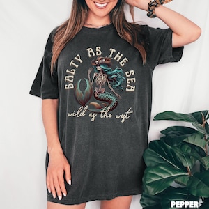 May include: Dark gray t-shirt featuring a skeleton mermaid graphic with turquoise hair and a cowboy hat. The text on the shirt reads "Salty as the Sea" and "Wild as the West".