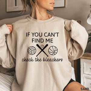 May include: Beige sweatshirt with the text "IF YOU CAN'T FIND ME check the bleachers". The design includes a volleyball, crossed baseball bats with a baseball, and a basketball. A casual, sporty design.