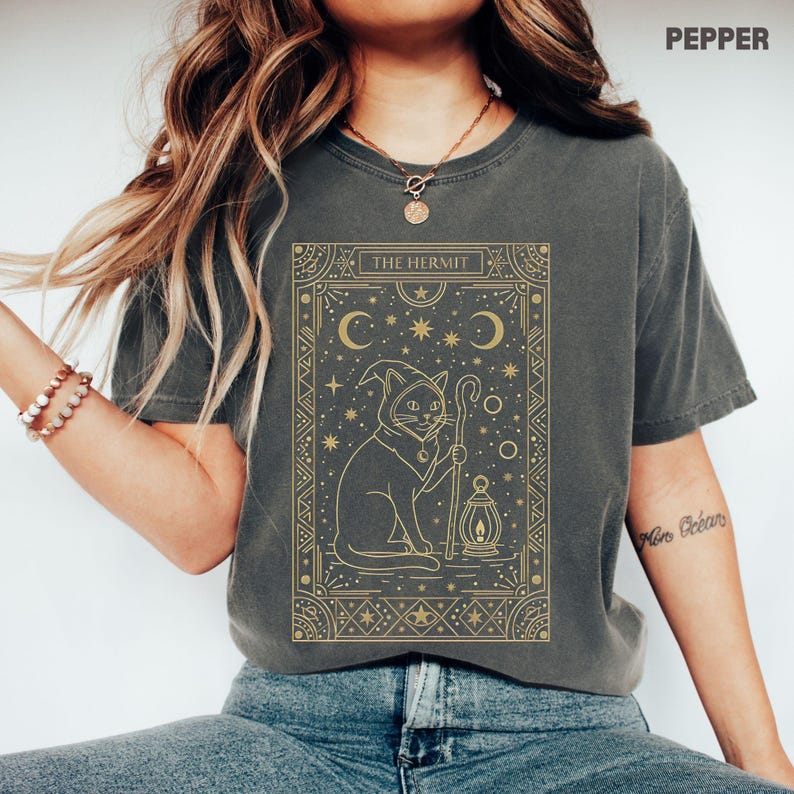 May include: A dark gray t-shirt featuring a gold-colored illustration of a cat in a hooded robe, holding a staff and lantern. The text "THE HERMIT" is at the top of the design, with crescent moons and stars. The shirt is a Pepper color.