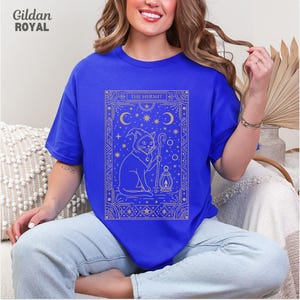 May include: Royal blue t-shirt featuring a gold illustration of a cat in a hooded cloak holding a staff and lantern, with the text "THE HERMIT". The design includes moons, stars, and decorative borders. The Gildan Royal brand is visible in the upper left corner.