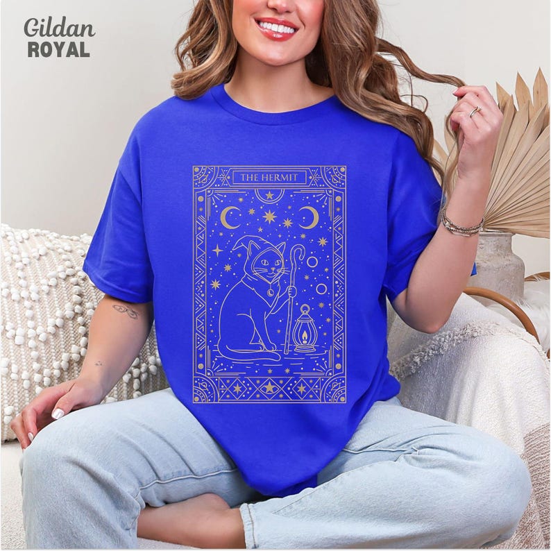 May include: Royal blue t-shirt featuring a gold illustration of a cat in a hooded cloak holding a staff and lantern, with the text "THE HERMIT". The design includes moons, stars, and decorative borders. The Gildan Royal brand is visible in the upper left corner.