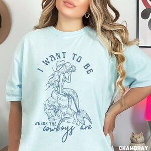 woman wearing a chambray shirt with a blue design showing a mermaid cowgirl and the phrase "i want to be where the cowboys are"