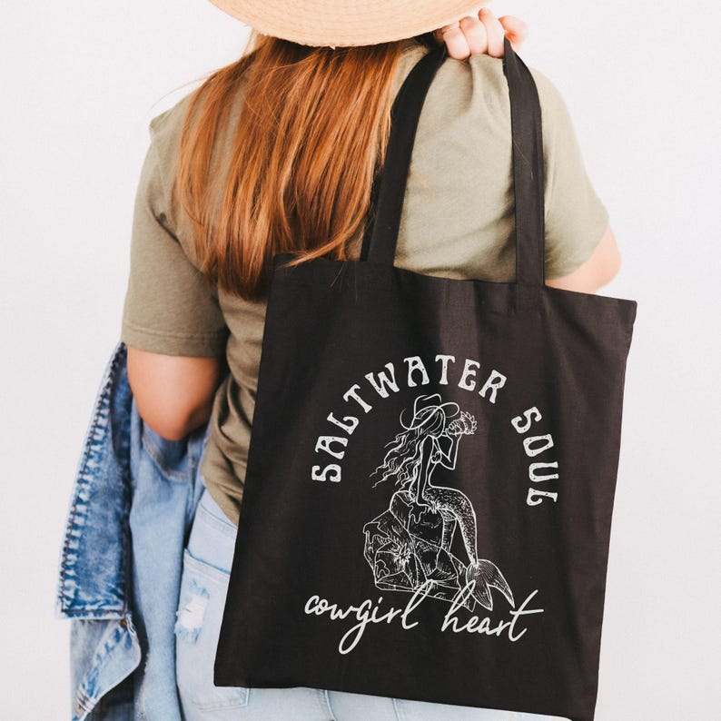 woman holding a black canvas tote bag with a blue design showing a mermaid cowgirl sitting on a rock and the phrase saltwater soul cowgirl heart