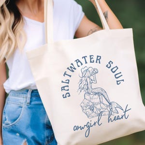 woman holding a beige canvas tote bag with a blue design showing a mermaid cowgirl sitting on a rock and the phrase saltwater soul cowgirl heart