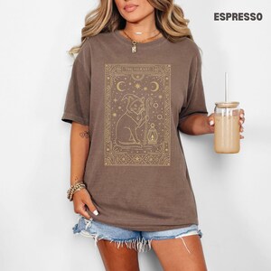 May include: Espresso-colored t-shirt featuring a gold illustration of a cat in a wizard's hat holding a staff and lantern, with the text "THE HERMIT". The design is framed with stars and crescent moons.