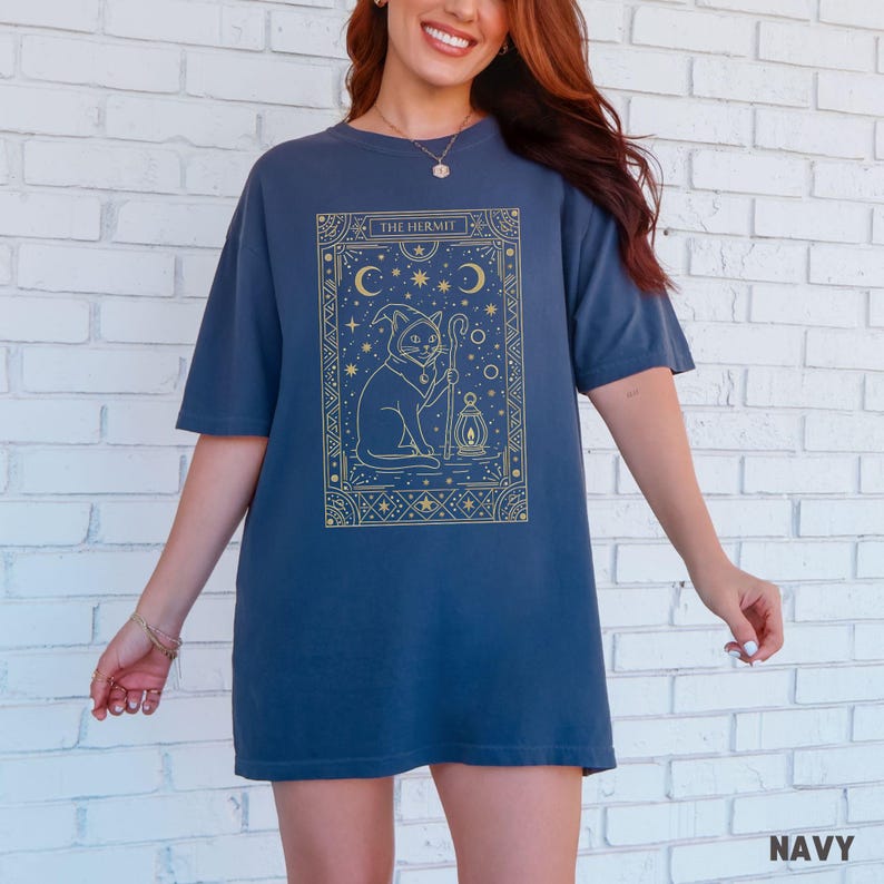 May include: Navy blue t-shirt featuring a gold-colored illustration of a cat in a hat holding a staff and lantern, with the text "THE HERMIT". The design includes moons, stars, and geometric patterns. The shirt is a comfortable, oversized fit.