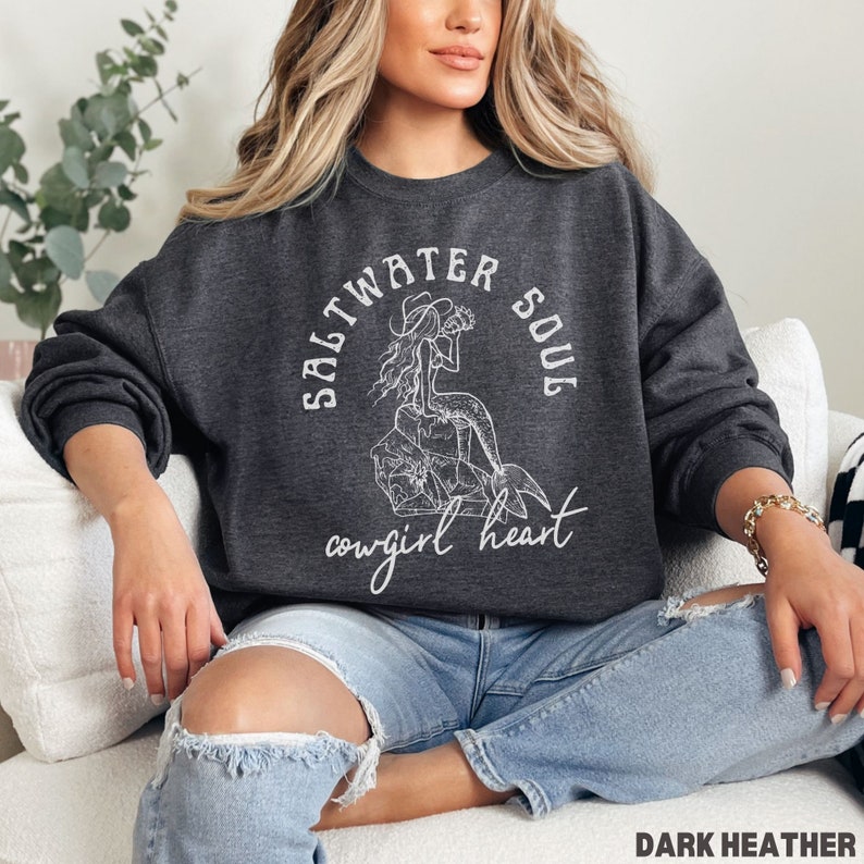 woman wearing a dark heather sweatshirt with a white design of a mermaid wearing a cowgirl hat and the phrase saltwater soul cowgirl heart