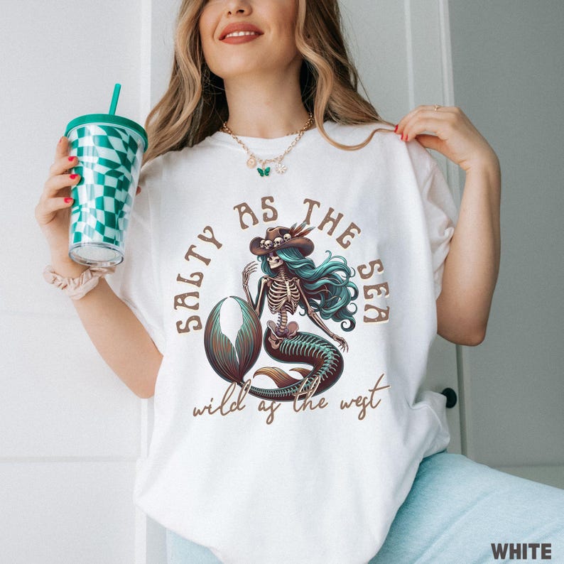 May include: White t-shirt with a graphic of a mermaid skeleton wearing a cowboy hat, with turquoise hair and a brown tail. The text reads "Salty as the Sea, Wild as the West". The shirt is a relaxed fit.