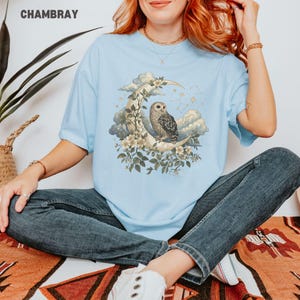 May include: Light blue t-shirt with a graphic of an owl perched on a crescent moon, surrounded by clouds, flowers, and stars. The word "CHAMBRAY" is printed at the top. The shirt is made of soft material.