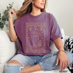 May include: Berry-colored t-shirt featuring a gold illustration of a cat in a wizard hat holding a staff and lantern, with the text "THE HERMIT". The design is framed with celestial elements. The shirt is paired with blue jeans.