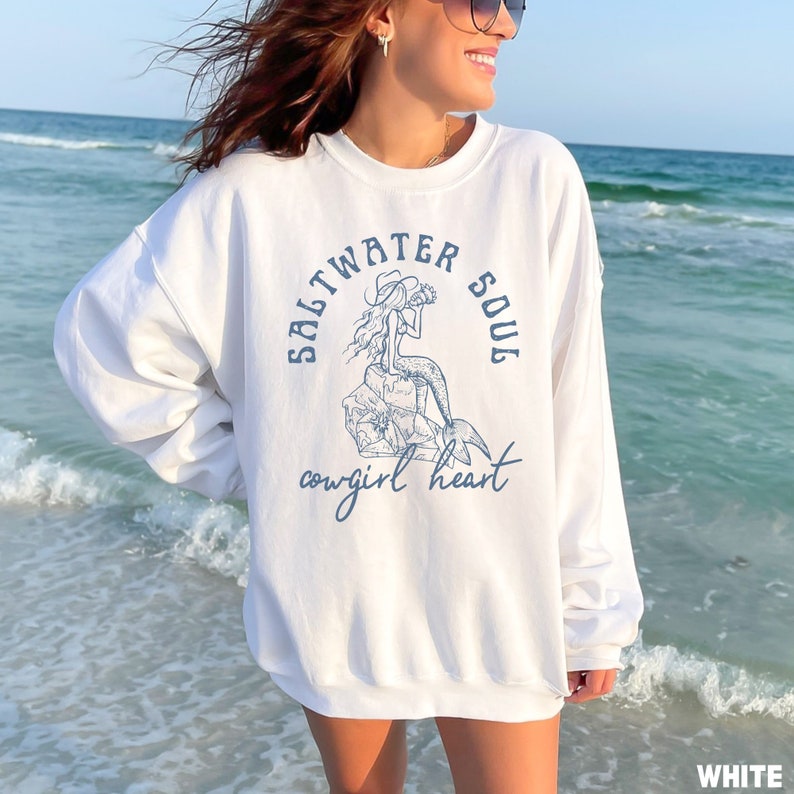 woman wearing a white sweatshirt with a blue design of a mermaid wearing a cowgirl hat and the phrase saltwater soul cowgirl heart