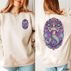 May include: Sand-colored sweatshirt featuring a mermaid graphic. The front has a small oval design with a mermaid tail. The back displays a larger, detailed mermaid illustration with a crown, surrounded by floral and geometric elements, in shades of blue, purple, and pink.