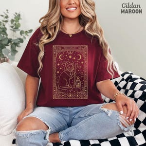 May include: Maroon t-shirt with a gold graphic of a cat wearing a wizard hat, holding a staff and lantern, with the text "THE HERMIT". The shirt is a Gildan brand.
