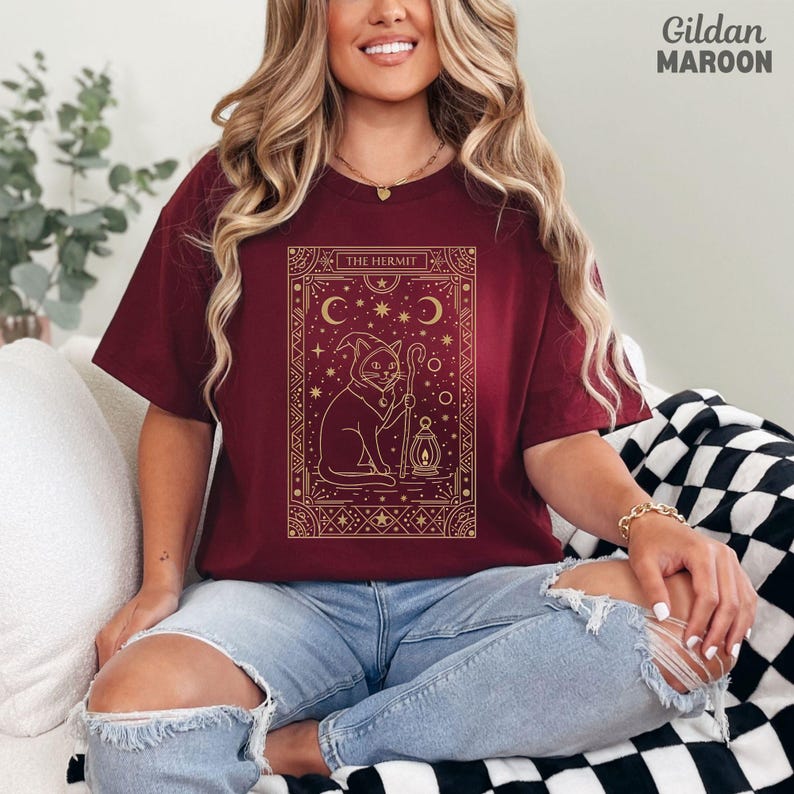 May include: Maroon t-shirt with a gold graphic of a cat wearing a wizard hat, holding a staff and lantern, with the text "THE HERMIT". The shirt is a Gildan brand.