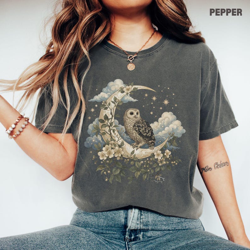 May include: A dark gray t-shirt featuring a whimsical design of an owl perched on a crescent moon, surrounded by clouds, flowers, and stars. The word "PEPPER" is printed at the top. The shirt is a comfortable, casual style.